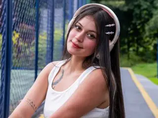LizzaRoss poses outdoors wearing a white tank top, showing her long dark hair and a tattoo on her upper arm.