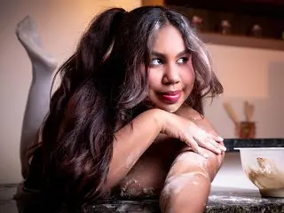 LizzieWilliams poses with long wavy hair, showing her bare shoulders and chest while leaning forward against a dark surface.