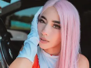 LolaTorress with long pink hair and a nose ring poses inside a car wearing a white top.