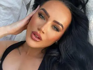 Lola_Valentina poses with her hand near her face, showing her long dark hair and heavy eye makeup.