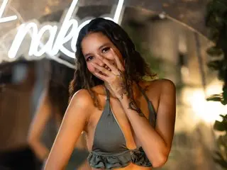LolaaDream poses with her hand over her mouth, wearing a low-cut olive green top that shows cleavage, with her wavy hair falling over her shoulders.