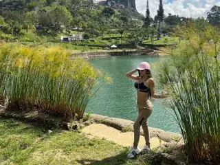 Lolaperrys poses outdoors in a black bikini top, showing her tanned skin and hair, while standing near a calm lake or pond surrounded by green grass.
