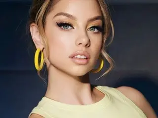 Young woman with blonde hair and yellow hoop earrings wearing a pale yellow top in a studio setting.