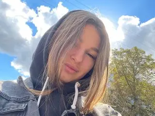 LonaSweet poses with long brown hair wearing a black hooded sweatshirt against a bright blue sky with white clouds.