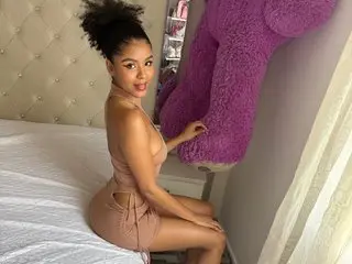 LoreyHamilton poses in a tan bodysuit, showing her cleavage and legs while sitting on a white bed against a light headboard.