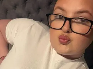 LottieRoseX wearing a white t-shirt and black glasses, posing with a pouty lip against a grey tufted headboard.