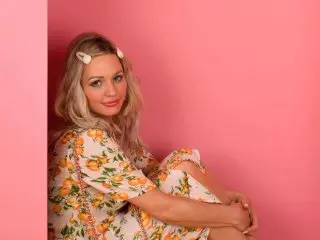 Lottie_Fun poses with long blonde hair against a pink background, wearing a floral print sundress.