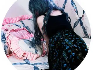 Young woman with long wavy blue hair wearing a black tank top and peacock print skirt on a pink floral bed.