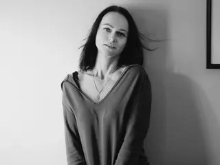 LoveMeINeed poses with long dark hair, wearing a deep V-neck top, looking directly at the camera against a plain light background.