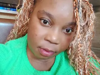 Close-up portrait of a woman with blonde braided hair wearing a bright green shirt in a casual indoor setting.