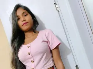 Lovelypriya poses against a white wall, wearing a pink short-sleeved button-up top with her long dark hair down.