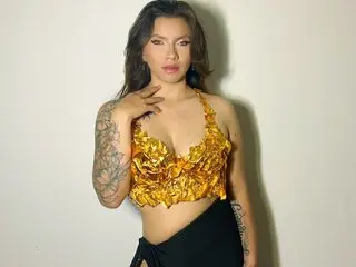 Lucia_s poses in a yellow ruffled crop top and black bottoms, showing off her tattoos and cleavage against a plain wall.