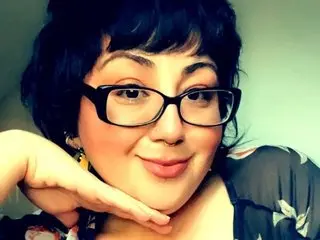 LuckLucy poses close-up with a smile, wearing black-rimmed glasses and a dark floral top, showing her dark hair and face.