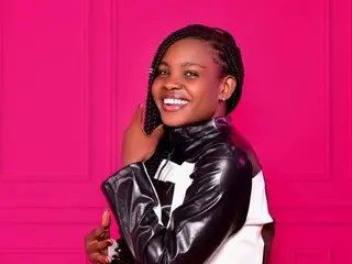 Lugwi69 smiles while wearing a black shiny leather jacket against a bright pink background.