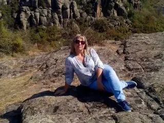 Lumo sits on a large rock outdoors wearing a grey shirt and blue jeans with sunglasses.