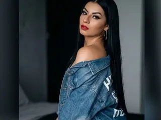 LunaCollinsss poses with long black hair, wearing an off-the-shoulder denim jacket that shows her bare shoulder and neck.