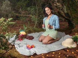 Smiling woman in a green floral dress and denim jacket enjoying a picnic in a sunny forest setting.