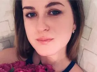 Luna_ria looks into the camera with long brown hair, wearing a dark top while holding a large bouquet of pink flowers near her chest.