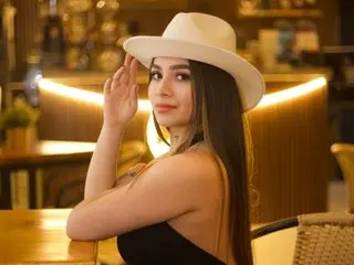 Young woman in a white wide-brimmed hat and black top smiling in a warm, dimly lit cafe setting.