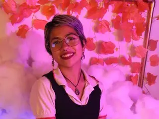 Lunarichy22 smiles while wearing a black and white top with glasses, set against a soft pink and red background.