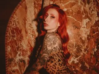 Lunaxluck poses with her long red hair and arm tattoos, wearing a black leopard print top against a warm marble background.