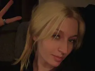 Blonde woman in a black outfit making a peace sign in a dimly lit indoor setting.