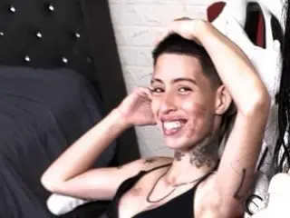 Smiling woman with short hair in a black tank top relaxing in a bedroom setting.