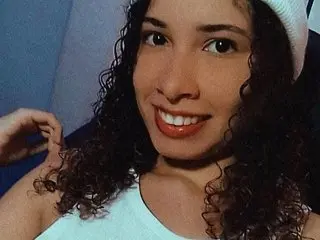 LupitaLopez smiles while looking at the camera, showing her long, dark curly hair and wearing a white tank top.