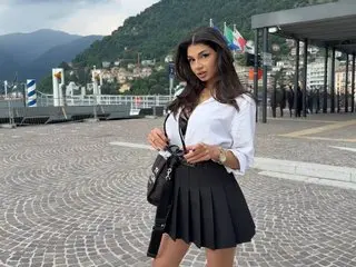 LuvCaprice poses outdoors in a white button-down shirt and a short black pleated skirt with her long dark hair down.