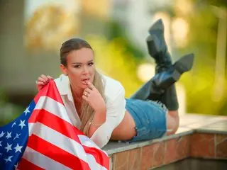 LuxenNoir — LuxenNoir lies on a ledge outdoors wearing a white shirt and denim shorts while holding an American flag.