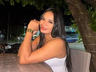 LyaSweetxx poses with a smile, wearing a white tank top and showing her long dark hair while resting her arm on a table outdoors at night.