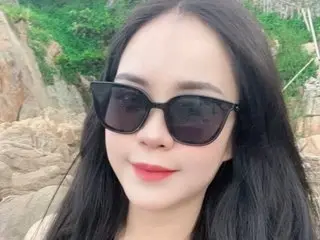 Smiling young woman with long dark hair wearing black sunglasses in a sunny outdoor rocky setting.