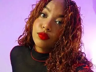 Young woman with curly red hair and red lipstick wearing a black turtleneck in soft purple lighting.