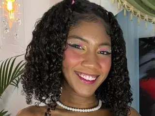 Smiling young woman with curly hair and colorful makeup wearing a pearl necklace in a bright indoor setting.