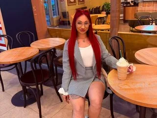MAIYRAxo poses in a grey blazer and matching skirt, looking at the camera with long red hair.