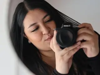 Young woman with long dark hair holding a professional DSLR camera in a bright indoor setting.