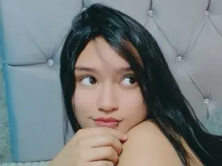 MEGAN_SOPHIA poses with her long dark hair, looking over her shoulder with a soft expression against a light blue tufted headboard.