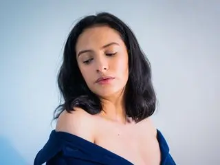 Melancholic young woman with dark hair wearing an off-the-shoulder navy blue top against a light blue background.