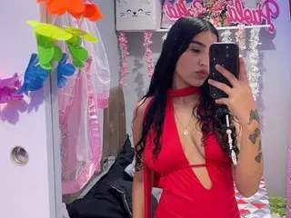 MIA_BROWN69 poses in a red top with thin straps, showing her long dark hair and cleavage against a light-colored wall background.