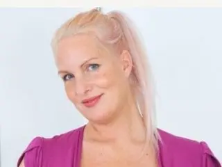 MJCoconut, a blonde woman, smiles at the camera while wearing a purple top against a plain white background.