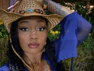 MZZZWETWET wearing a brown straw hat and blue top, showing her face and long dark hair in an outdoor setting.