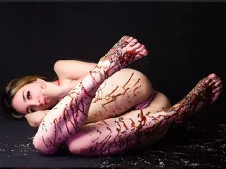 Maaiia lies on a black surface, her body and legs covered in thick, dripping chocolate sauce.