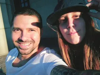 MacaThompson smiles next to a man in a white t-shirt, showing her long brown hair and face in a close-up selfie.