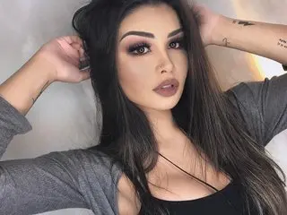Young woman with long dark hair and glamorous makeup wearing a grey top in a soft, moody indoor setting.