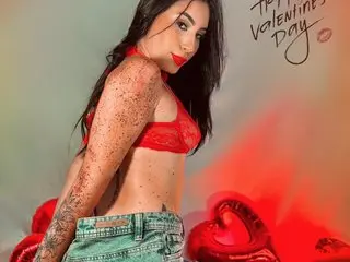 MaddiKayy poses in a red lace bra and denim shorts, showing off her long dark hair and bare back against a red heart background.
