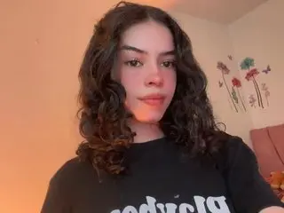 Young woman with curly brown hair wearing a black graphic t-shirt in a warm, cozy bedroom setting.