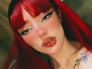 MaddyMorgan with bright red hair and winged eyeliner looks into the camera with a tongue piercing.