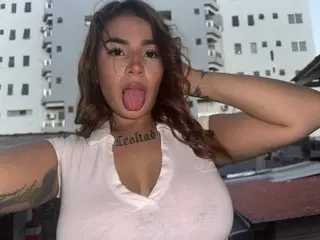 Maddy_Aniston poses in a white tank top, showing her cleavage and long brown hair while sticking her tongue out at the camera.