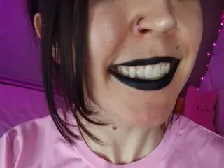 MadysonMinxxx shows her face and smile while wearing a pink t-shirt and dark lipstick, with dark hair framing her face.