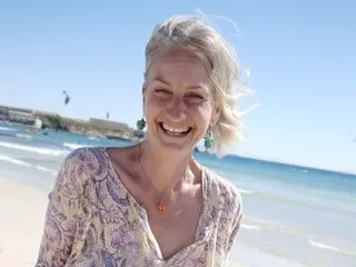 MagicMia smiles with her blonde hair loose, wearing a light patterned blouse while standing outdoors near the beach and ocean.